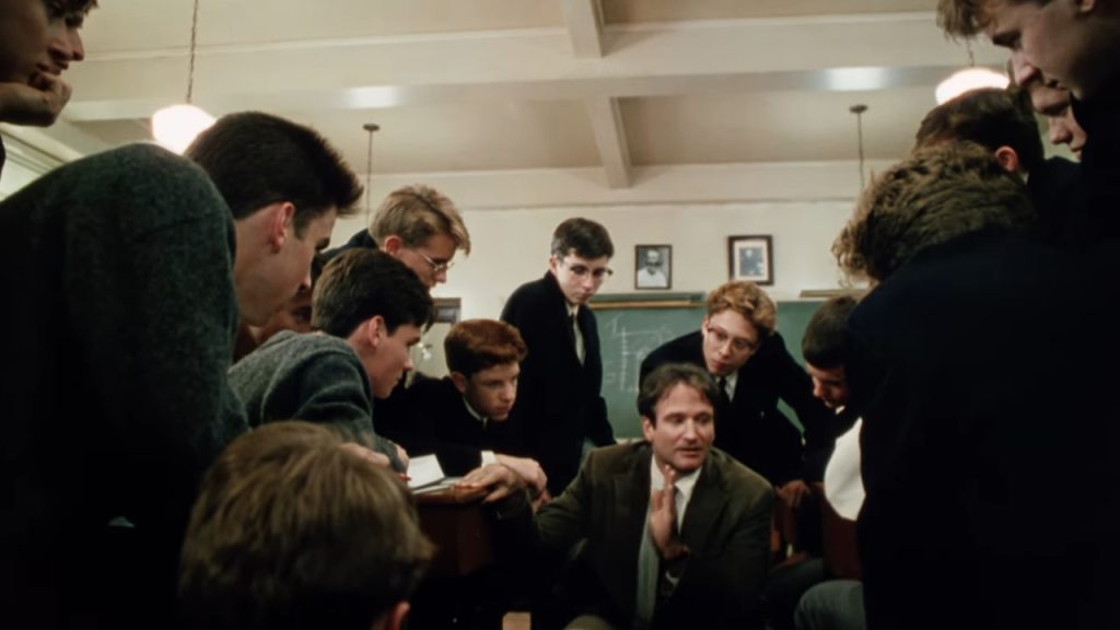 A still from Dead Poets Society. In a classroom, a man in an olive suit crouches among a circle of uniformed boys that listen to him with interest.