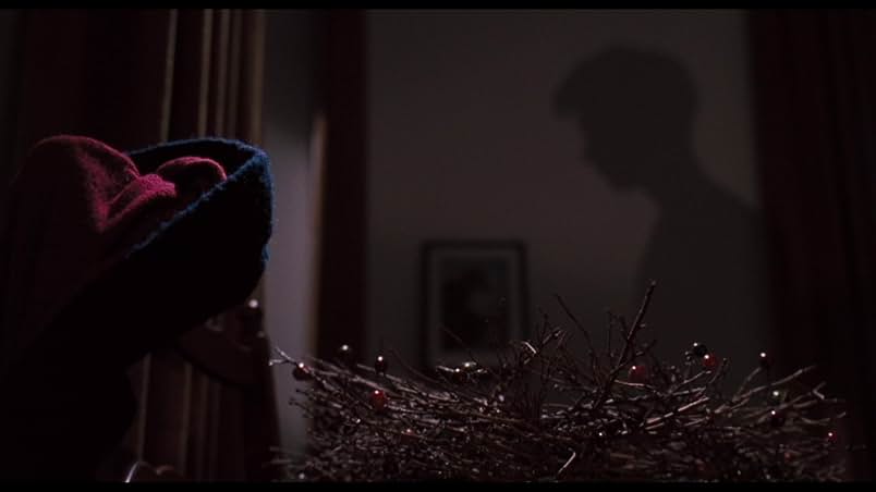 A still from Dead Poets Society. In a dimly lit room, a wreath made of brown sticks and red berries sits beside blue scarf and red gloves. On the wall in the background is a man's shadow.