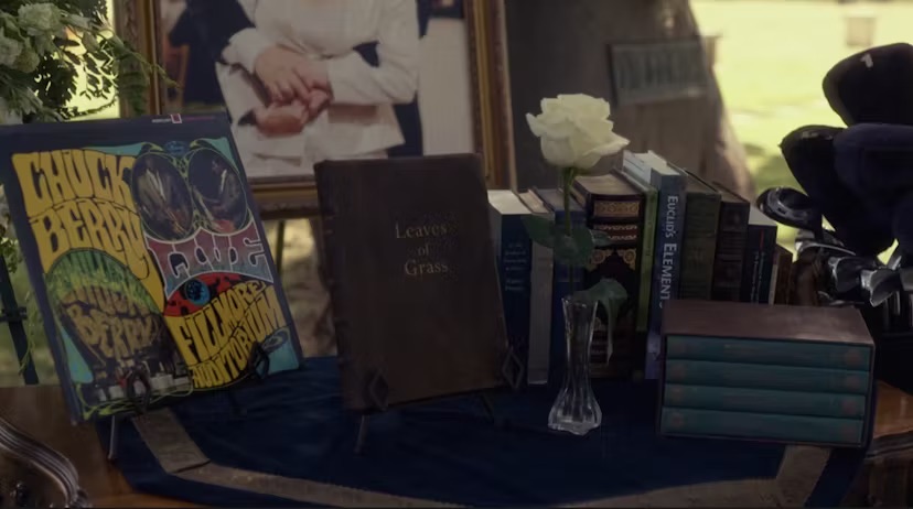 A still from Gilmore Girls: A Year in the Life. On a dark wood table with a blue velvet cloth are a collection of items: a Chuck Berry record, a copy of Leaves of Grass, a white vase in a jar, and multiple stacks of books. Leaning against the table are golf cllubs.