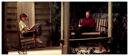 A still from The Notebook. A young man sits on a rocking chair outside reading from a book. Sitting opposite from him on a porch swing is an older man in a red shirt.