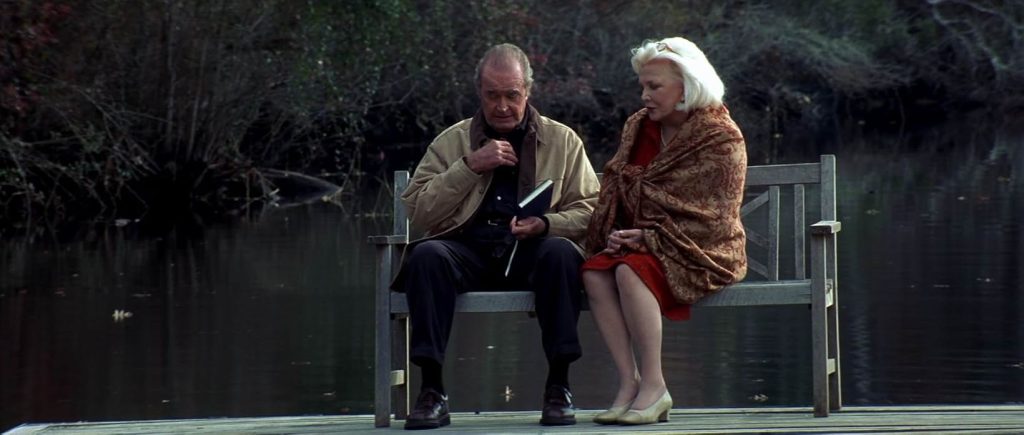 A still from The Notebook. An older man in a brown jacket and dark pants sits on a white bench beside an older woman wearing a red dress and brown shawl. Behind them is a lake.