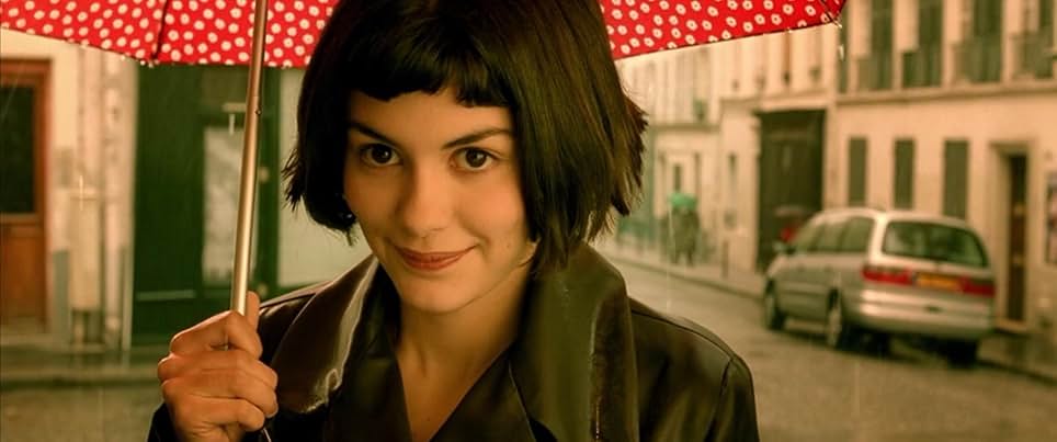 A still from Amélie. A woman with short dark hair wears a dark jacket and smiles while holding a red patterned umbrella.