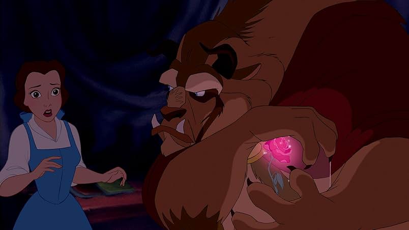 A still from the 1991 version of Beauty and the Beast. A young woman in a blue dress with a white undershirt looks on in shock at a large beast figure with brown fur, a lion-like snout, and dark horns. He clamps his paws protectively around a glowing pink rose in a glass display case. The beast glares at the young woman menacingly.