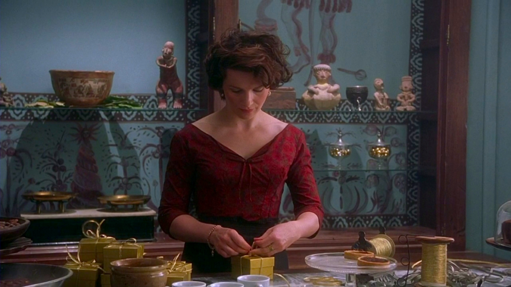 A woman in a red top stands in a chocolaterie wrapping boxes in gold paper and ribbon. The wall behind her is blue and hand painted with brown Mayan drawings.