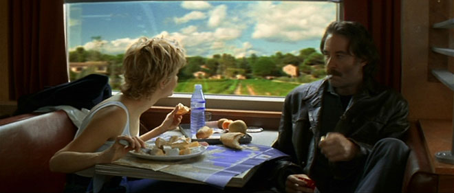 A still from French Kiss. A blonde woman in a white tank top leans over a plate of cheese and looks out the window at a gorgeous country view. Opposite her at the booth is a man wearing a dark jacket with a cigarette in his mouth.