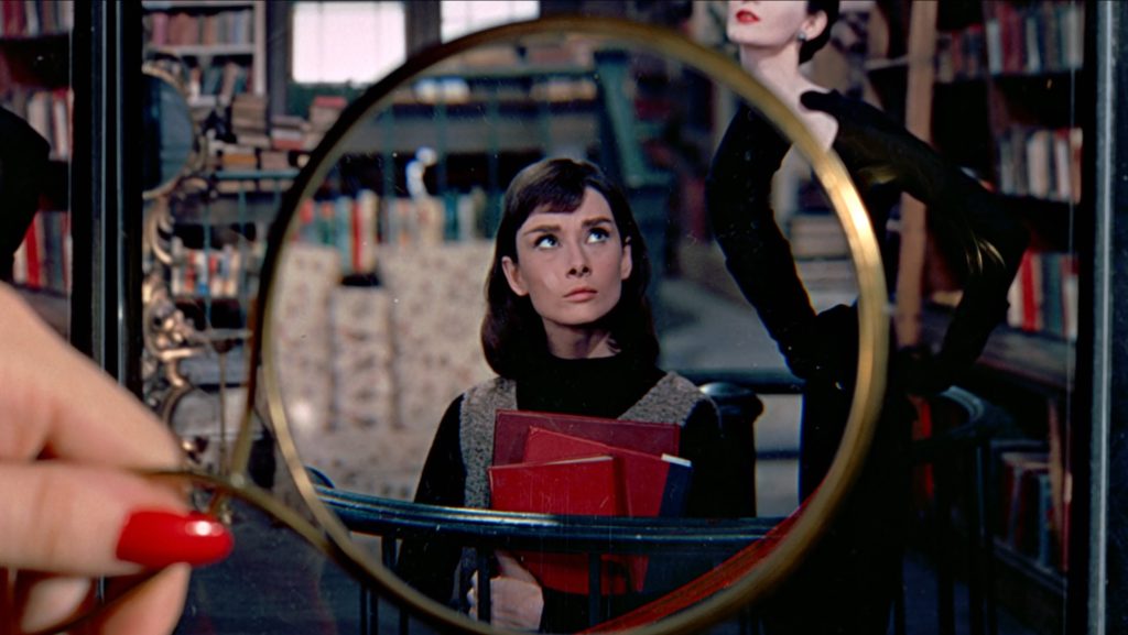 A still from Funny Face. A young woman with brown hair holds a stack of red books in a bookshop and looks up at a posing model. A hang with red fingernails holds a gold magnifying glass in front of the frame that emphasizes the woman holding the books.