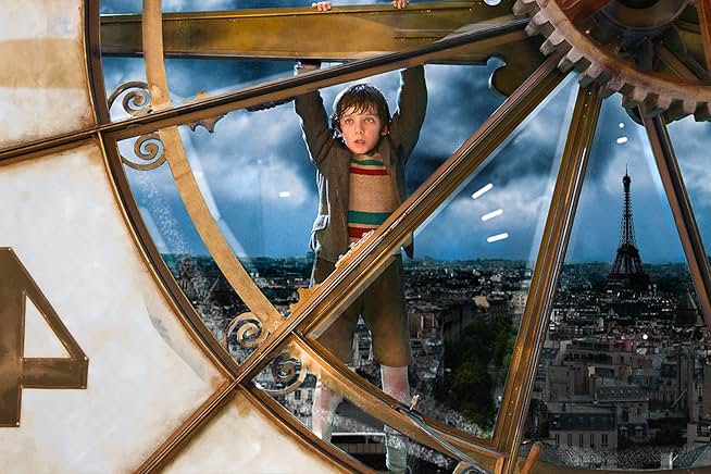 A still from Hugo. A boy in a brown jacket and trousers with a tan, green, and red striped sweater hangs onto the outer edge of a golden clock face. Through the clear glass is a view of the Paris skyline, complete with the Eiffel Tower.