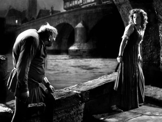 A black-and-white still from the 1939 version of The Hunchback of Notre Dame. A hunchbacked man follows a woman wearing a long dress. She looks over her shoulder at him. Behind them is a river.