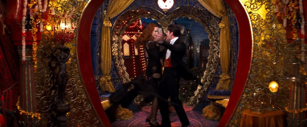 A still from Moulin Rouge!. A redheaded woman in black lingerie dances with a man in black tuxedo with a red vest. They are in a gilded room decorated with yellow, red, and blue accents. The entryway they dance in is shaped like a heart.