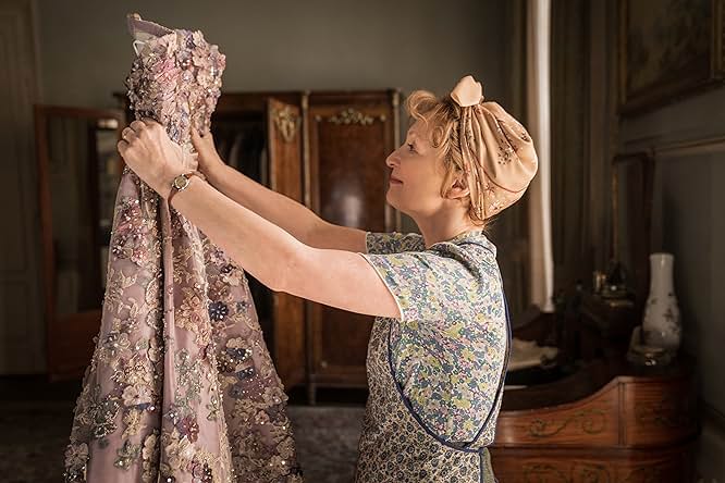 A still from Mrs. Harris Goes to Paris. A woman wearing a blue floral dress with matching work apron and a pink headscarf holds up and admires a purple gown covered in multicolored intricate beading work.