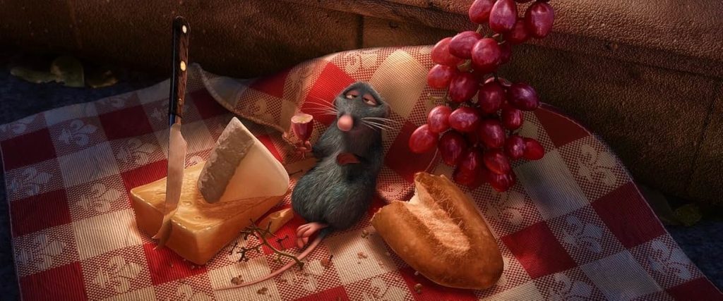 A still from Ratatouille. A gray rat sits on a red and white checkered cloth with a fleur de lis pattern surrounded by food. There are red grapes, part of a baguette and two different types of cheese with a knife embedded in one of the blocks.