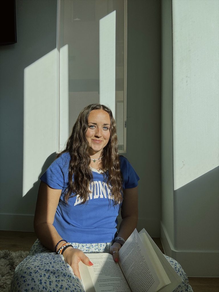 Bookstagrammer Madisyn sitting on the floor with her back against the wall and a mirror. She sits basked in sunlight with a sweet smile on her face, her blue eyes lit up by the sun, wearing a blue shirt that say "KINDNESS" in white, and has an open book on her lap.