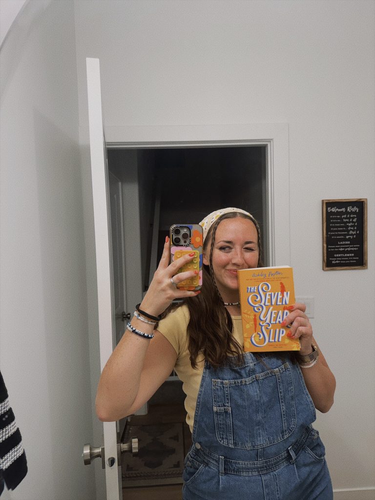 Bookstagrammer Madisyn in blue overalls, a yellow top, and a white and yellow bandana with two braids peeking out the front taking a mirror selfie with her flowery phone. She is displaying the yellow romance book cover for The Seven Year Slip by Ashley Poston.