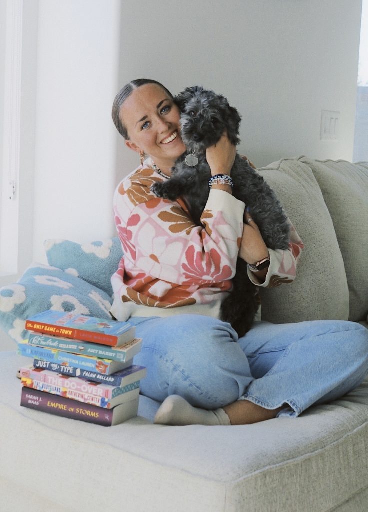 Bookstagrammer Madisyn sitting on a tan couch in a pink and brown flowery sweater, lifting up her little black dog to her cheek. Next to her is a stack of seven books; visible titles include: Empire of Storms by Sarah J.Maas, The Do-Over by Lynn Painter, Just My Type by Falon Ballard
