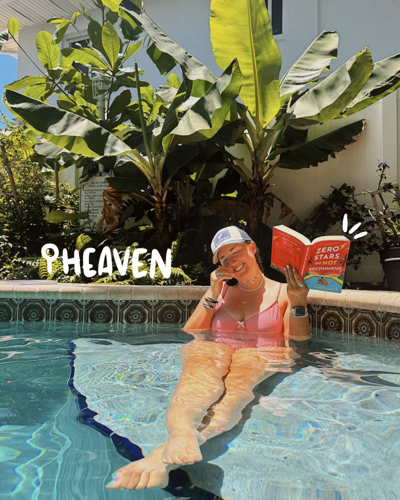 Bookstagrammer Madisyn usrrounded by lush green plants while laughing in a pool. In a red and white plaid bathing suit, a blue and white hat, and holding open Zero Stars Do Not ERecommend by M.J. Wassmer, Madisyn soaks in the sun,