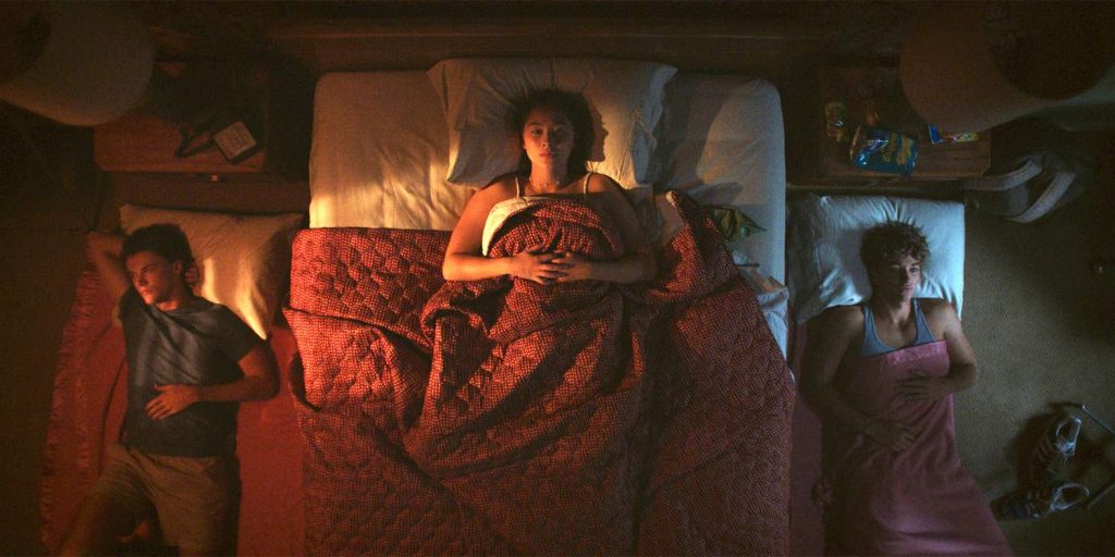 Belly (center) lays on the bed between Conrad (left) and Jeremiah (right) who are on the floor on either side of the bed