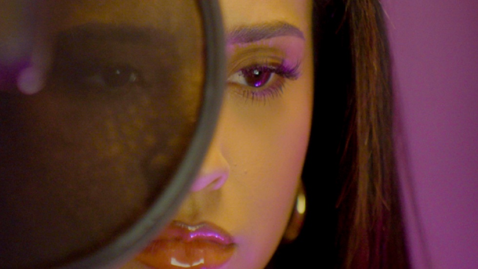 Rebbeca film still. A closeup of Becky G's face with a solemn expression, partially obscured by a round mic.