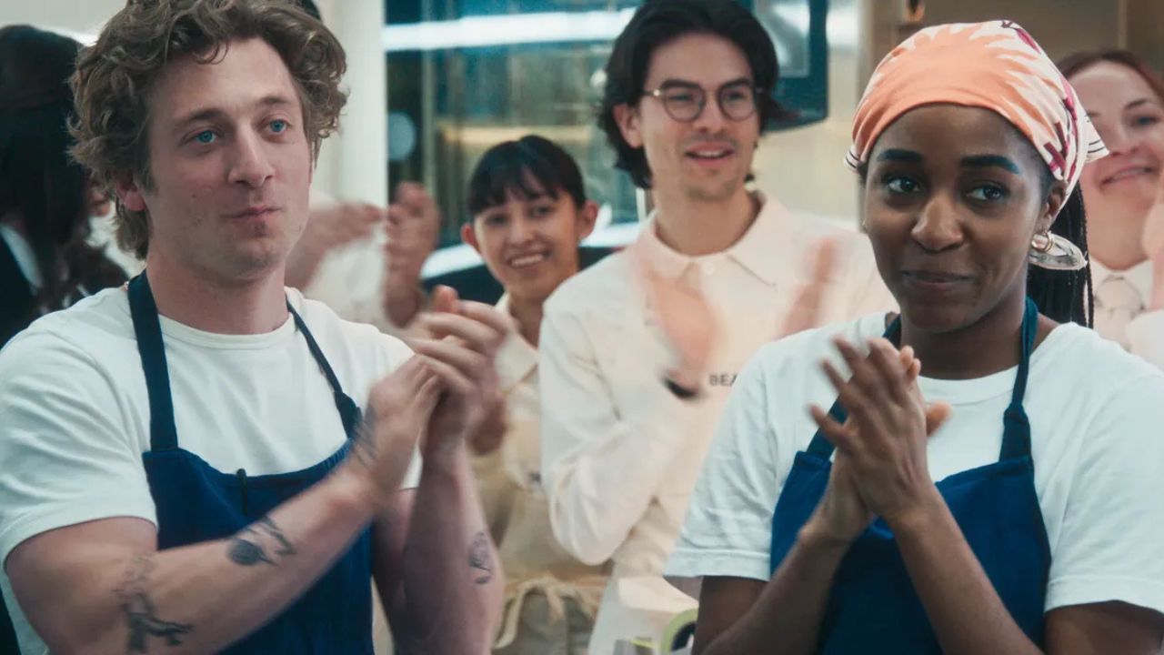 The Bear, season four episode 9. Carmy (Jeremy Allen White) and Syd (Ayo Edebiri) stand in the kitchen with proud smiles on their face as they clap their hands; the kitchen is full with other workers clapping in support. Both Carm and Syd are in white tshirts with dark blue aprons on