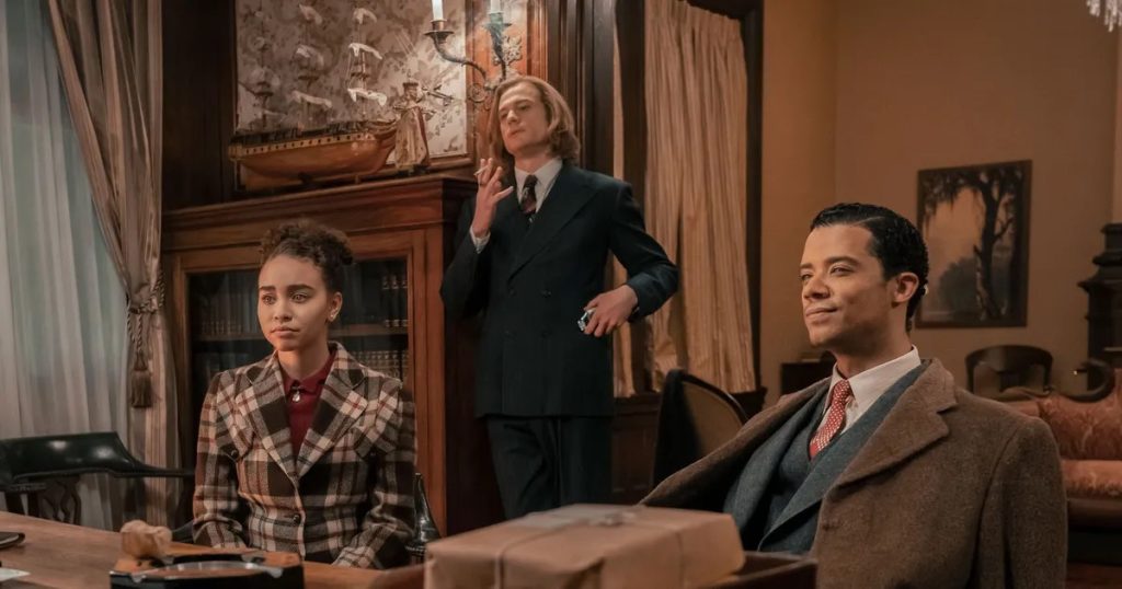 From left to right: Claudia de Pointe du Lac de Lioncourt (played by Bailey Bass), Lestat de Lioncourt (played by Sam Reid), and Louis de Pointe du Lac (played by Jacob Anderson) sit in a richly decorated room in the AMC 2022 series Interview with the Vampire. Claudia wears a plaid jacket, Lestat stands smoking a cigarette in a sharp suit, and Louis sits with a small smile in a brown overcoat.