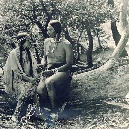 A black-and-white still from the 1920 film The Daughter of Dawn. A Native woman and man in traditional dress sit on a log in a forest.
