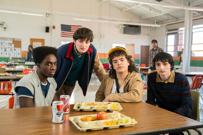 Image of Noah Schnapp (Will Byers), Finn Wolfhard (Mike Wheeler), Gaten Matarazzo (Dustin Henderson), and Caleb McLaughlin (Lucas Sinclair) sitting in a school cafeteria. They are all looking towards the camera with serious faces.