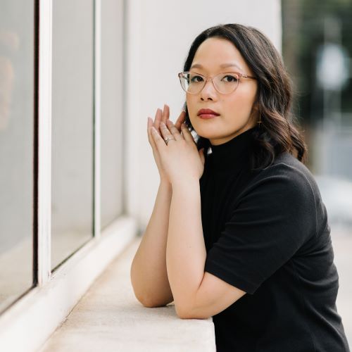 Trang Thanh Tran, a light-skinned Asian person wearing glasses and a black shirt, rests their elbows on a windowsill as they turn to face the viewer.