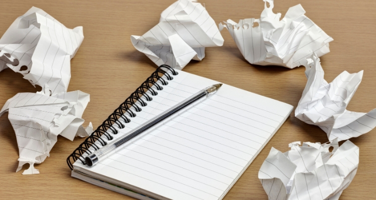 A blank notebook page with a black pen resting on it; both sit on a wooden surface surrounded by more crumpled-up pages of the notebook.