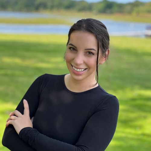 Headshot of Ellie K. Wilde. She has her arm crossed and is smiling at the camera. Her dark hair is in an updo, and she is wearing a long-sleeved black top. The background is out of focus, but it shows a grassy field and a body of water..