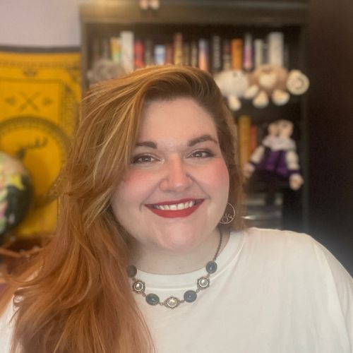 Headshot of Katie Chandler. She is pale-skinned, has long brunette hair, and is wearing a white shirt and silver and blue necklace. The background is out of focus, but it shows a bookshelf full of books and artwork on the left side.