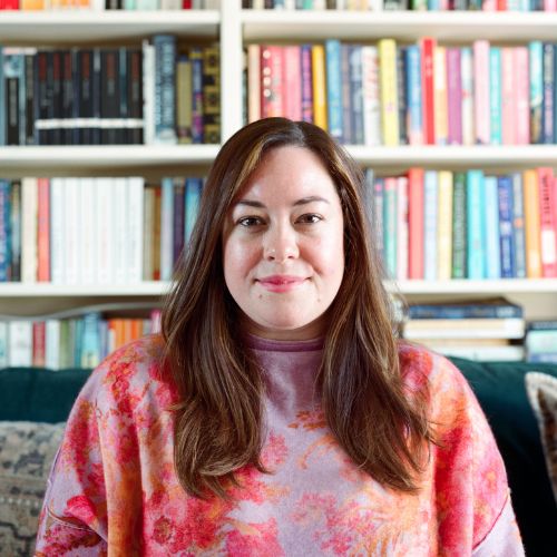 Headshot of Laura Wood. She is pale-skinned, has long brunette hair, and is wearing a tie-dye jumper. The background is out of focus, but it shows a bookshelf full of books.