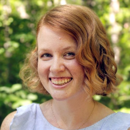 Headshot of Martha Waters. She is pale-skinned, has ginger short hair, and is wearing a blue and white blouse. The background is out of focus, but seems to be full of plants.