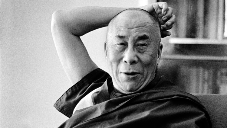 Black and white photo of the 14th Dalai Lama smiling and holding his hand behind his head.
