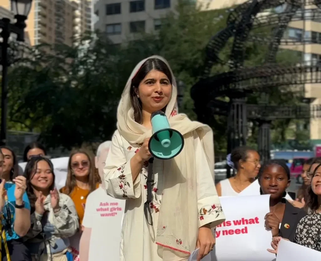 Malala stands in front of a smiling crowd and carries a blue megaphone.