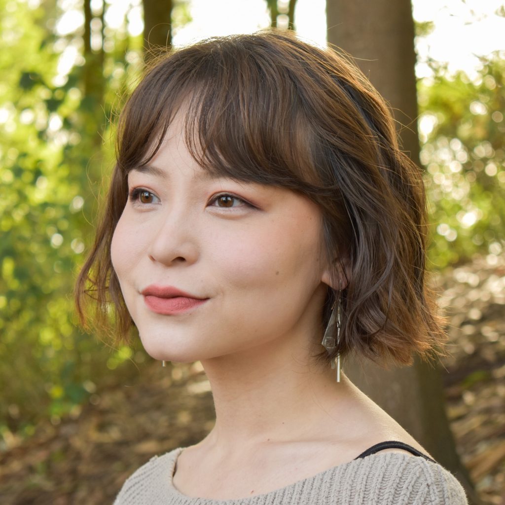 Angie Kang, a smiling East Asian light-skinned woman with chin-length brown wavy hair and brown eyes, wearing a beige sweater and posing in a wooded area.