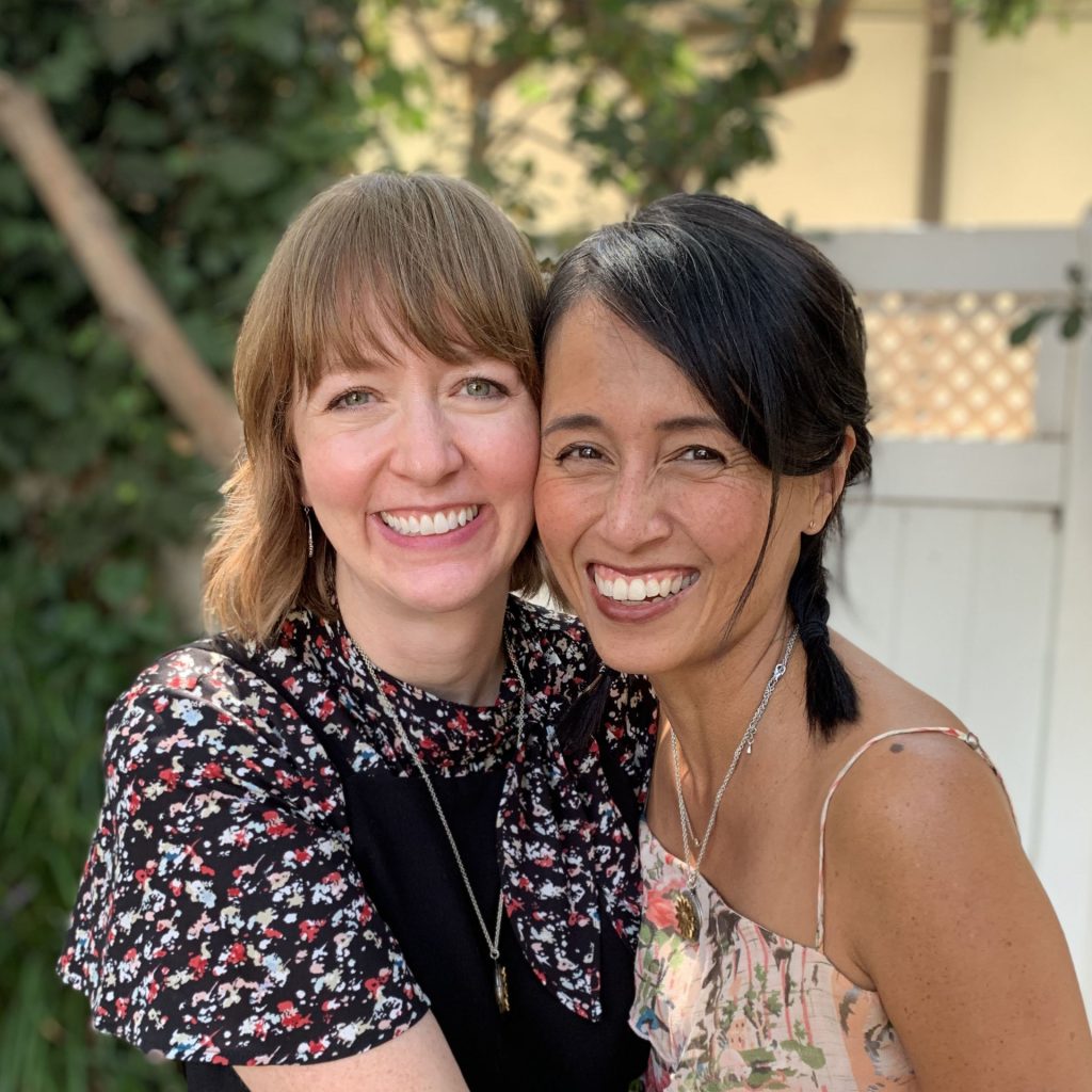 Shannon Hale (left) and LeUyen Pham (right) embracing and smiling for the camera. Shannon is a light-skinned woman with shoulder-length, light brown, wavy hear and green eyes, wearing a black and dark floral dress. LeUyen is a tan-skinned woman of East Asian descent with straight, braided black hair and brown eyes, wearing a pink floral dress.