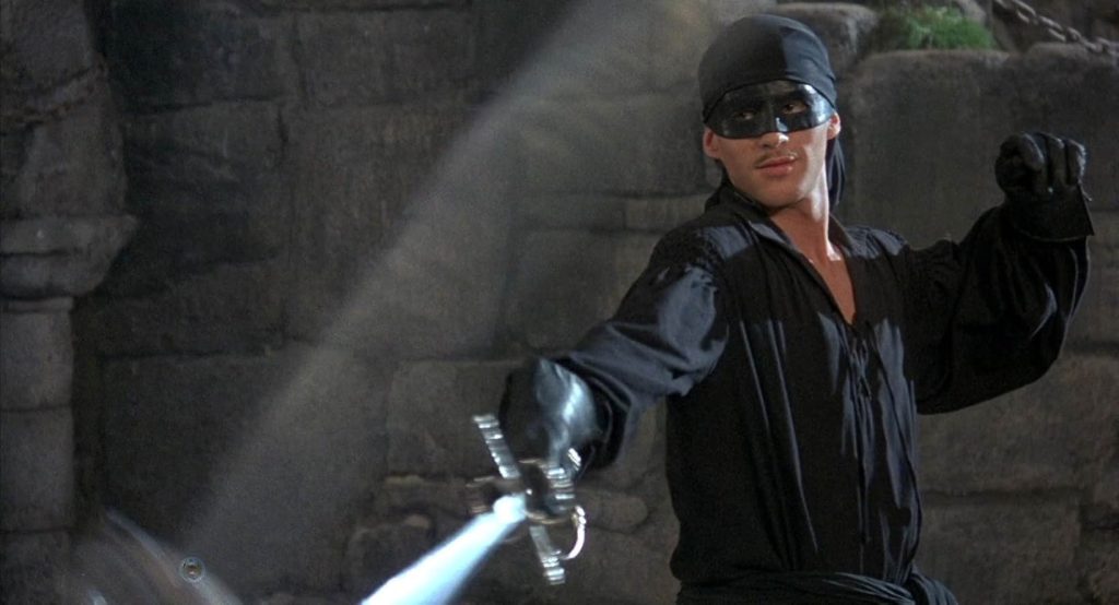 A still from the 1987 movie The Princess Bride. A man in a black blouse, a black mask, and black gloves slashes his sword forward in combat. Behind him is a gray rock wall.