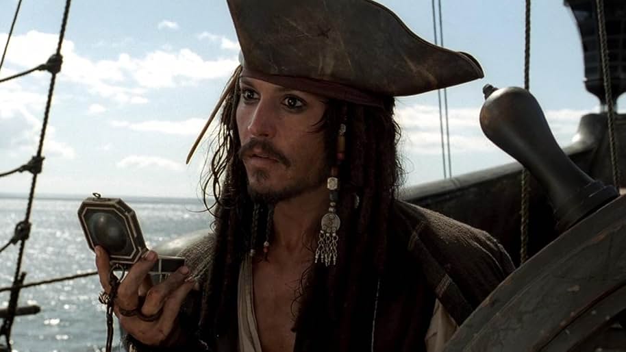 A still from the 2003 movie Pirates of the Caribbean: The Curse of the Black Pearl. A man with long dark hair woven with beads wears a brown leather tricorne cap and holds a black compass in his hand. He stands behind a ship wheel on the ocean and stares off into the distance.