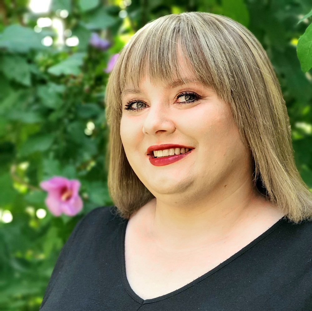 Headshot of Adrienne Rivera looking at the camera and smiling. She has shoulder-length blonde hair with bangs, red lipstick, and a black shirt on. In the background, there is a green bush with pink flowers.