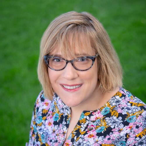 Author Claire Forrest, a lign-skinned woman with blond hair, wears glasses and a floral shirt smiles at the camera while outside.
