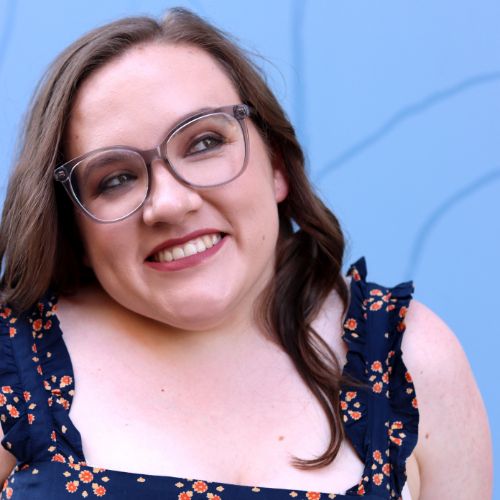 Author Gretchen Schreiber, a light-skinned woman with dark brown hair wearing glasses and a floral blouse smiles before a camera, her eyes looking to the side.