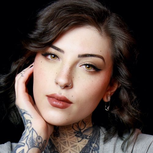Author McKenzie Catron, a light-skinned woman with dark brown hair and tattoos on her hand, arm, and neck, looks pensively into the camera.