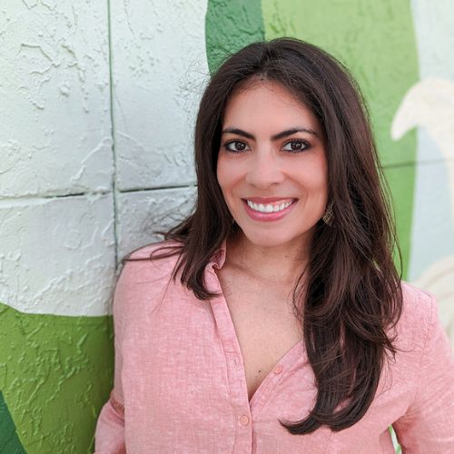 Author Natalia Sylvester, a Latinx woman with dark brown hair smiles sweetly to the camera wearing a pink linen shirt leaning against a mural painted brick wall.