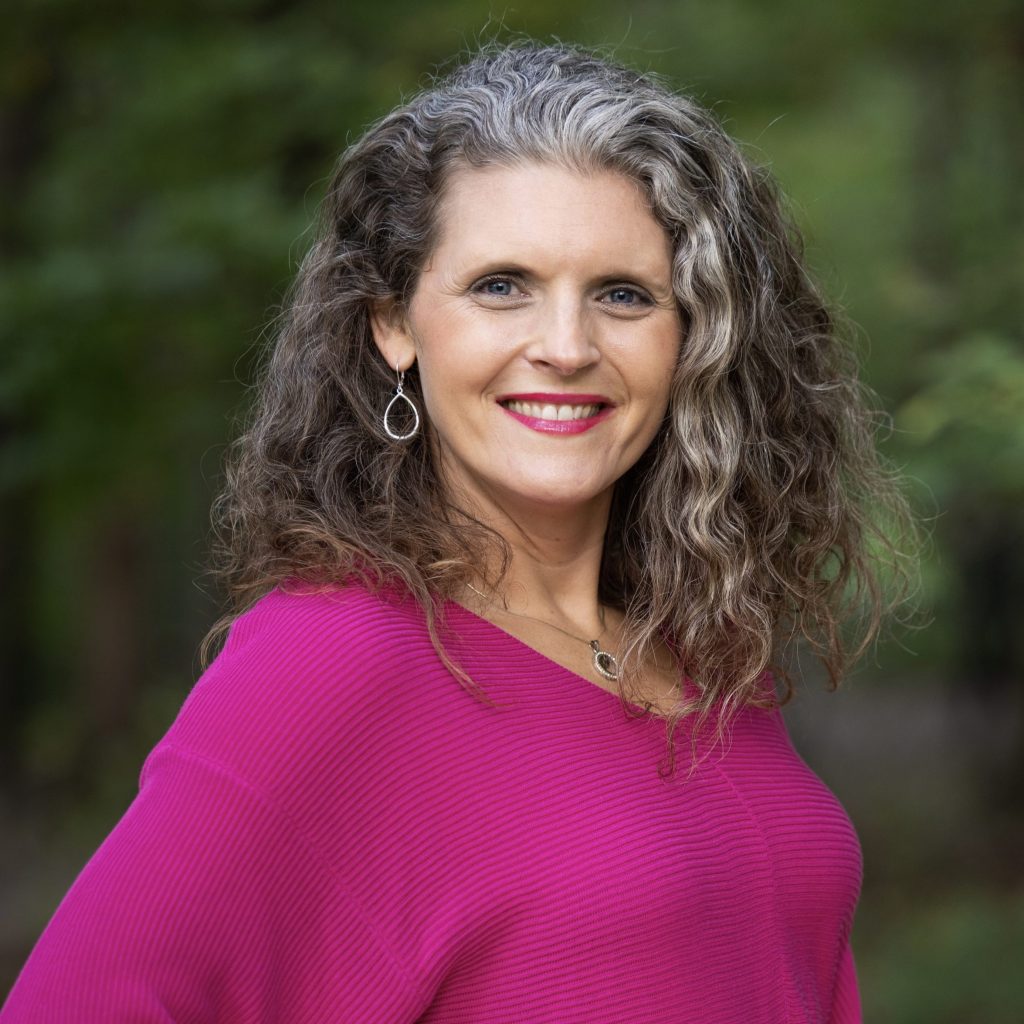 Lindsay Bonilla, a smiling light-skinned woman with shoulder-length salt-and-pepper hair and blue eyes, wearing a pink sweater and lipstick while posing in a wooded, outdoor area.