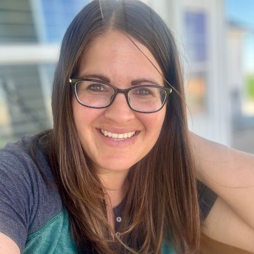 Romance author Cindy Steel, a light skinned brunette woman smiles at the camera wearing glasses, and a Henley, while outside.