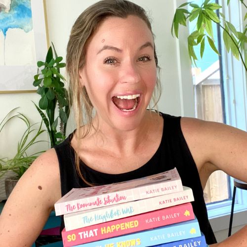 Romance author Katie Bailey, a light skinned blonde woman smiles widely while holding her stack of books.