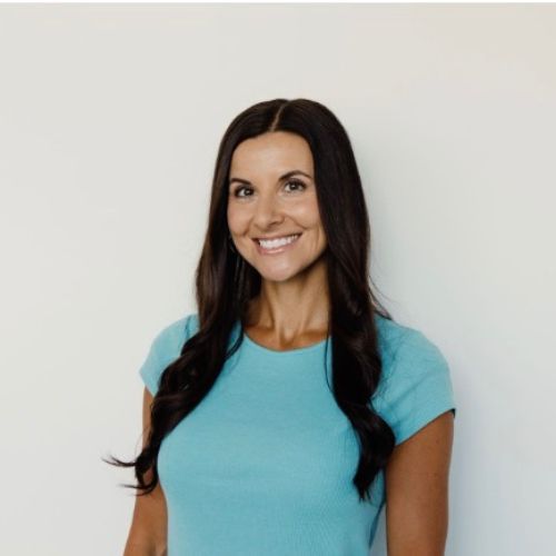 Romance author Larissa Hommes, a light skinned dark brunette woman smile at the camera wearing a light blue t-shirt before a beige wall.