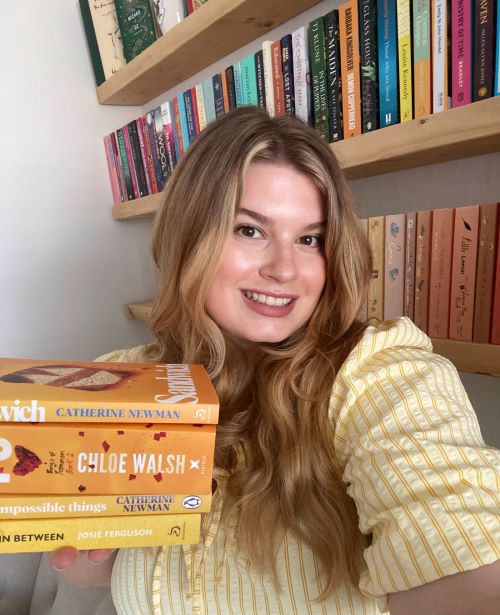 Clo from @theseasidebookclub, a light-skinned woman with long, wavy blonde hair and a long-sleeved white and yellow top, holds a stack of four books with yellow covers while smiling at the viewer. Behind her are shelves full of books; the wall is white.