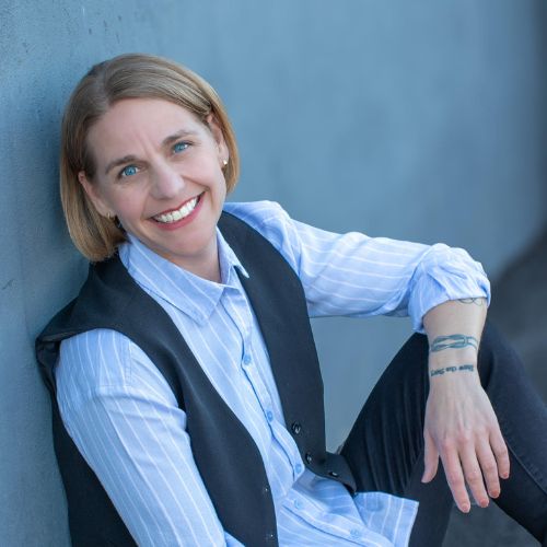 Author Court Stevens, a light skinned blond haired blue eyed woman leans against a wall wearing a vested suit smiling happily at the camera