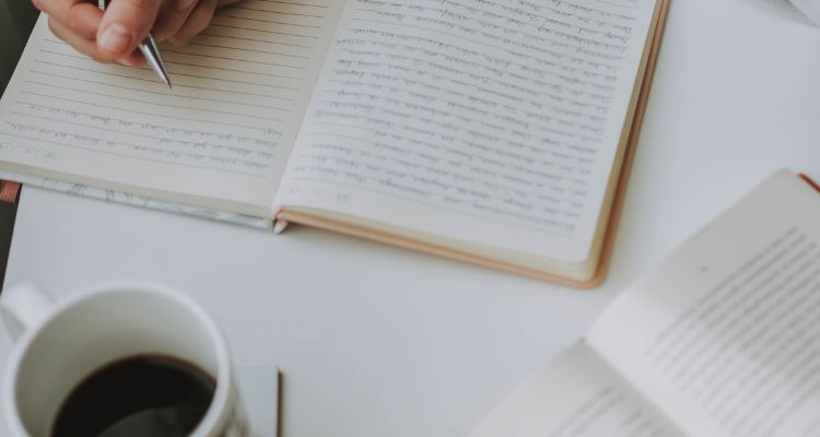 Depicts a person's hand writing in a journal with a cup of coffee and an open book.