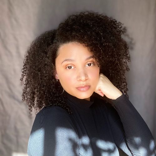 Headshot of Kaylnn Bayron in a black long-sleeve shirt staring at the camera. She has dark brown curly hair and is wearing silver hoop earrings.The background is grey.